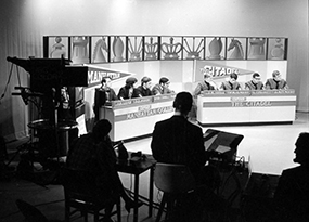 4 Manhattan College male students and 4 Citadel male students are seated at 2 tables in a television studio preparing to film the quiz show competition the College Bowl in March 1970.